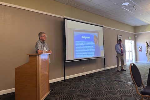A man on the left of a projector screen leans on a podium; another man stands to the right of a projector screen.